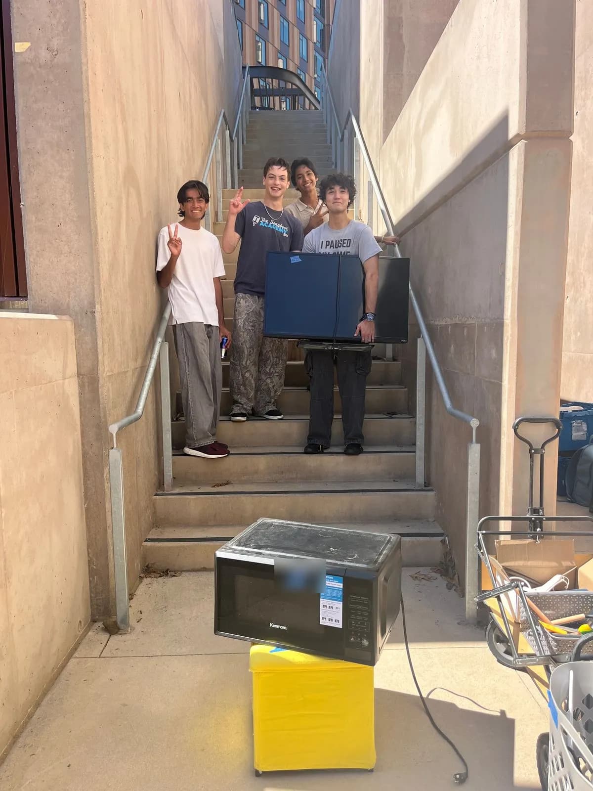 Four students on a stairway holding a TV, with a microwave on a storage ottoman in front of them