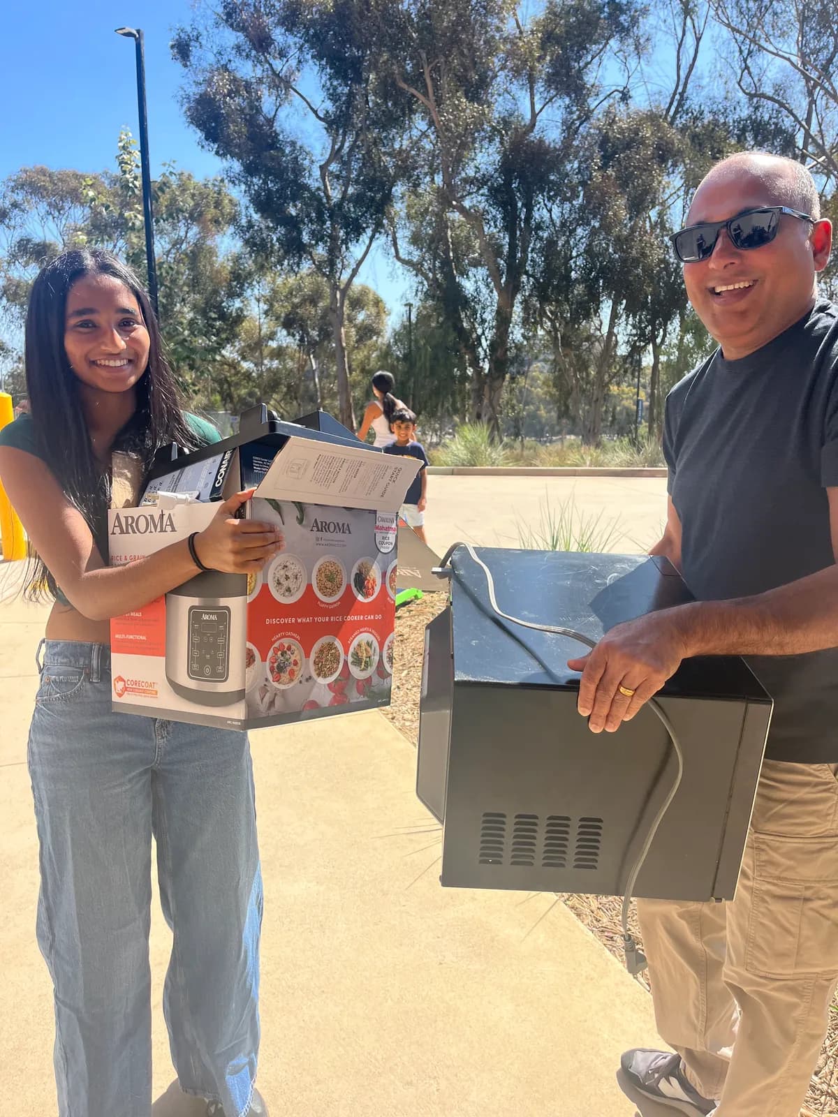 A student holding a boxed rice cooker beside a parent carrying a microwave at pickup