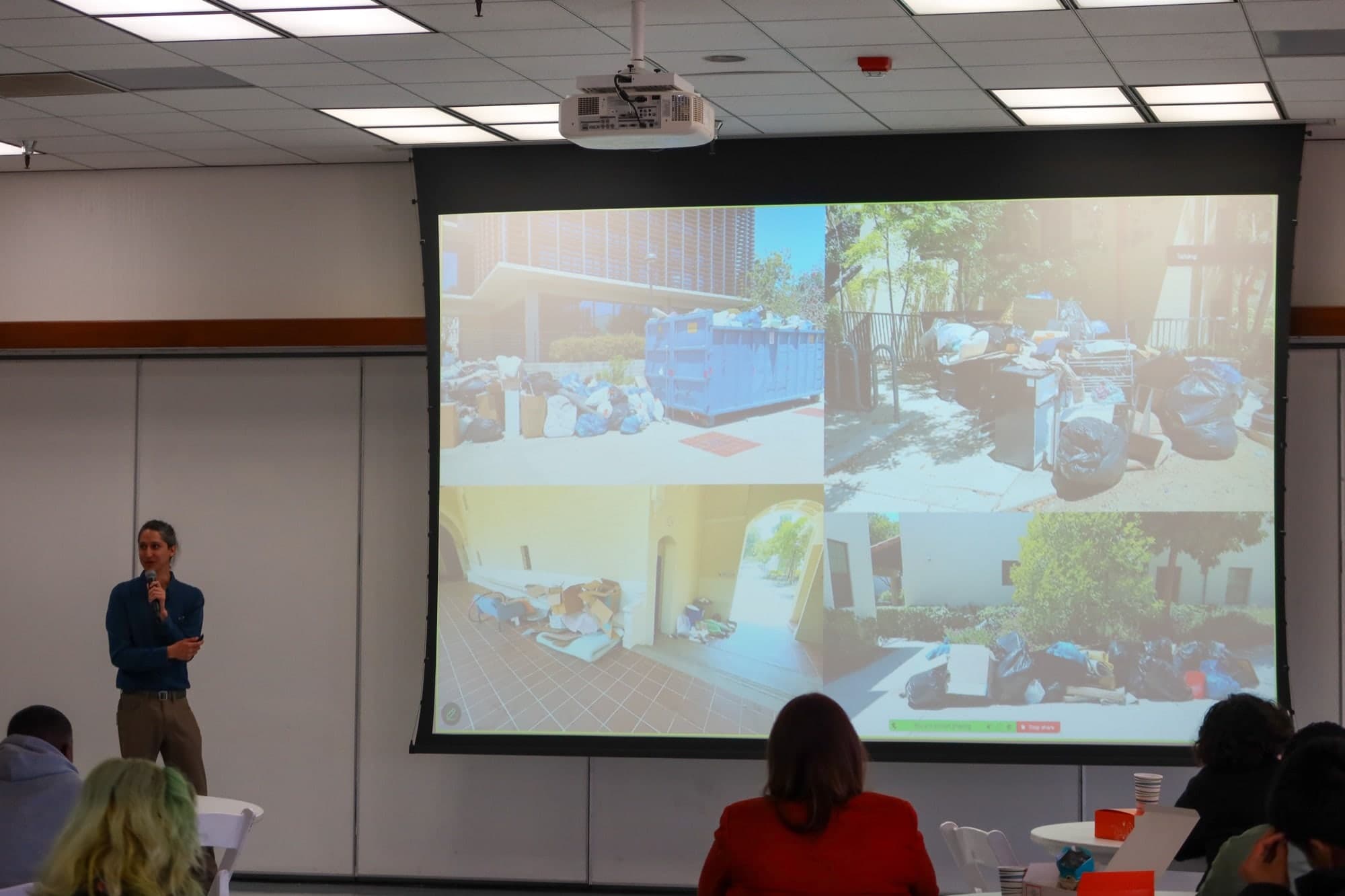 A speaker stands beside a projected grid of four photographs showing overflowing dumpsters, bagged trash, mattresses and discarded furniture piled outside residence halls