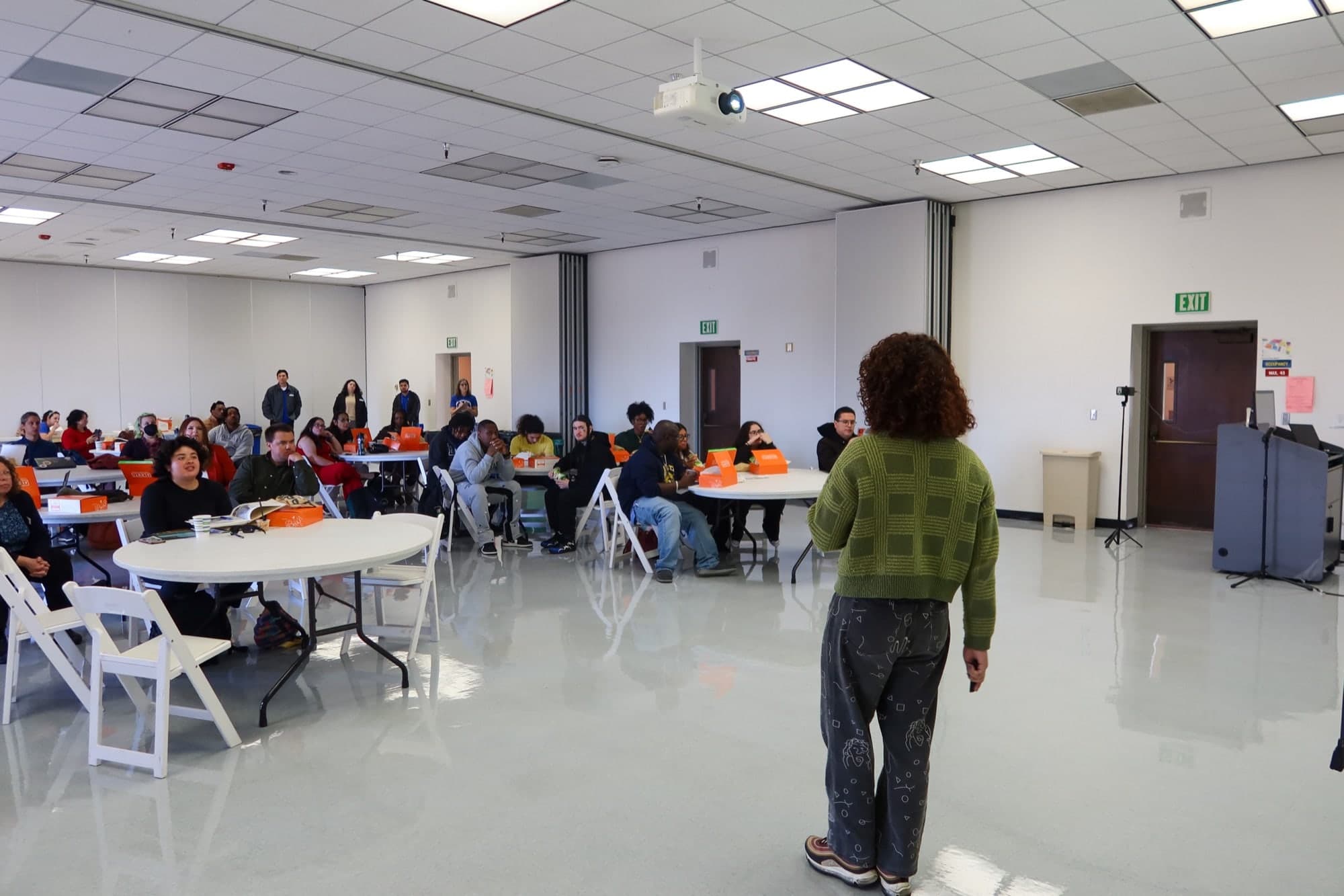 A wide view of a college multipurpose room with about thirty seated attendees at round tables, boxed lunches in front of them, facing a presenter in a green cardigan standing with their back to the camera
