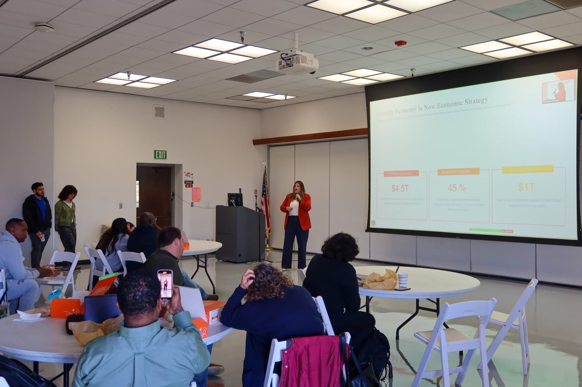 Lindsey Rowell, in a red blazer, holds a microphone in front of a projected slide reading Circular Economy Is Now Economic Strategy, with three statistics displayed beneath it