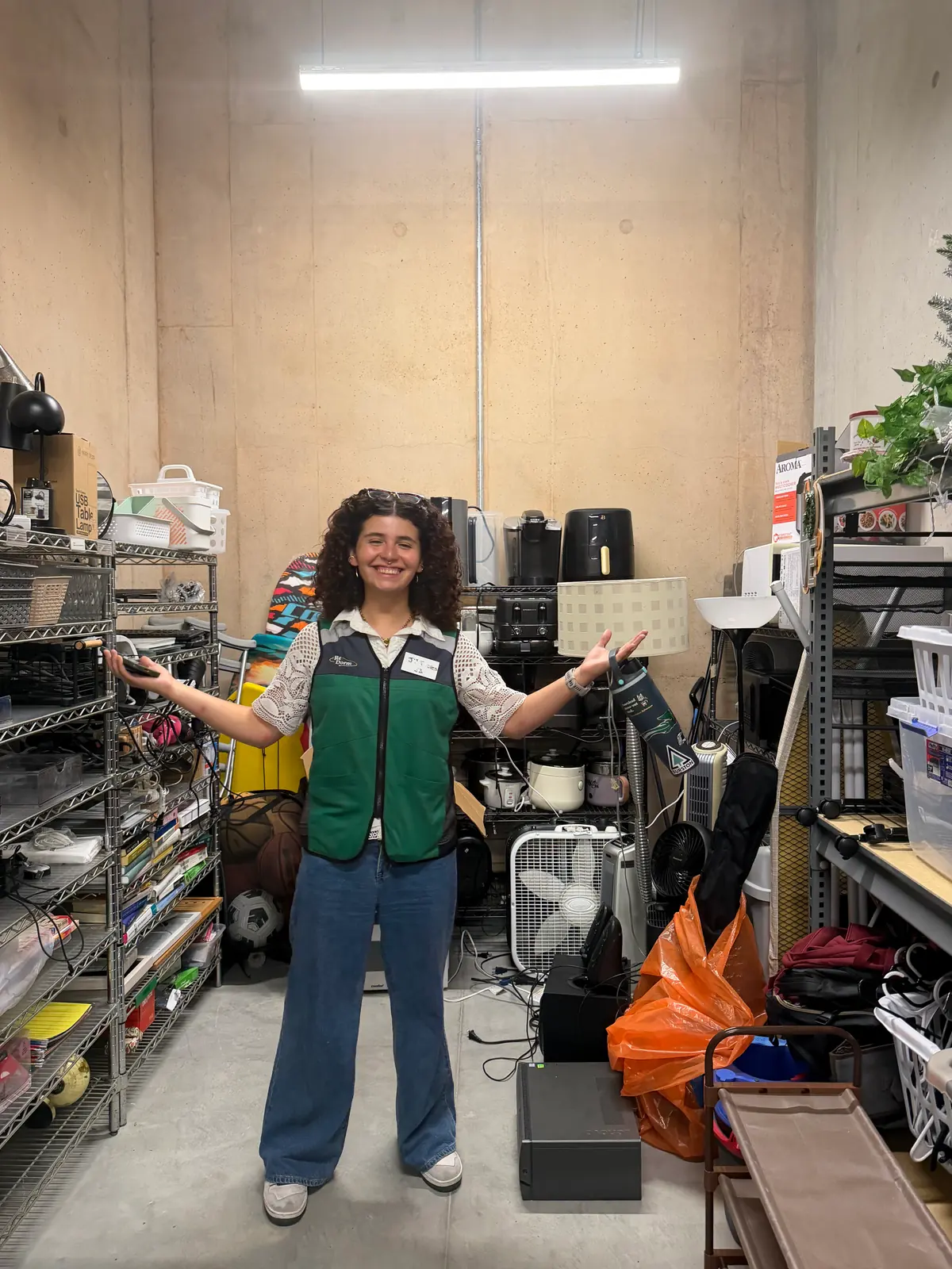A person in a green ReDorm vest standing with arms outstretched in a narrow storage room, surrounded by shelves of lamps, small appliances, fans, and household goods