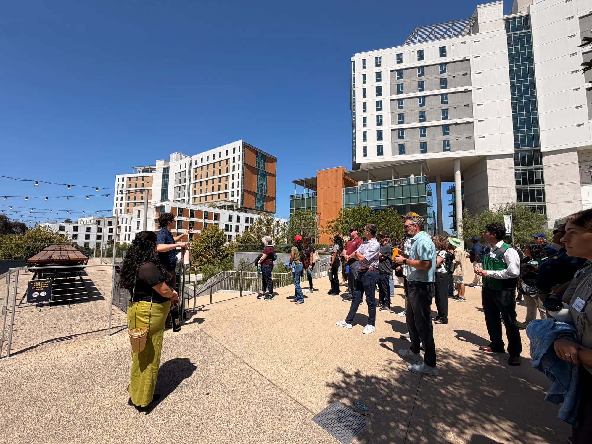 A large group of workshop attendees standing outdoors on a plaza between UC San Diego residence halls, listening to a presenter