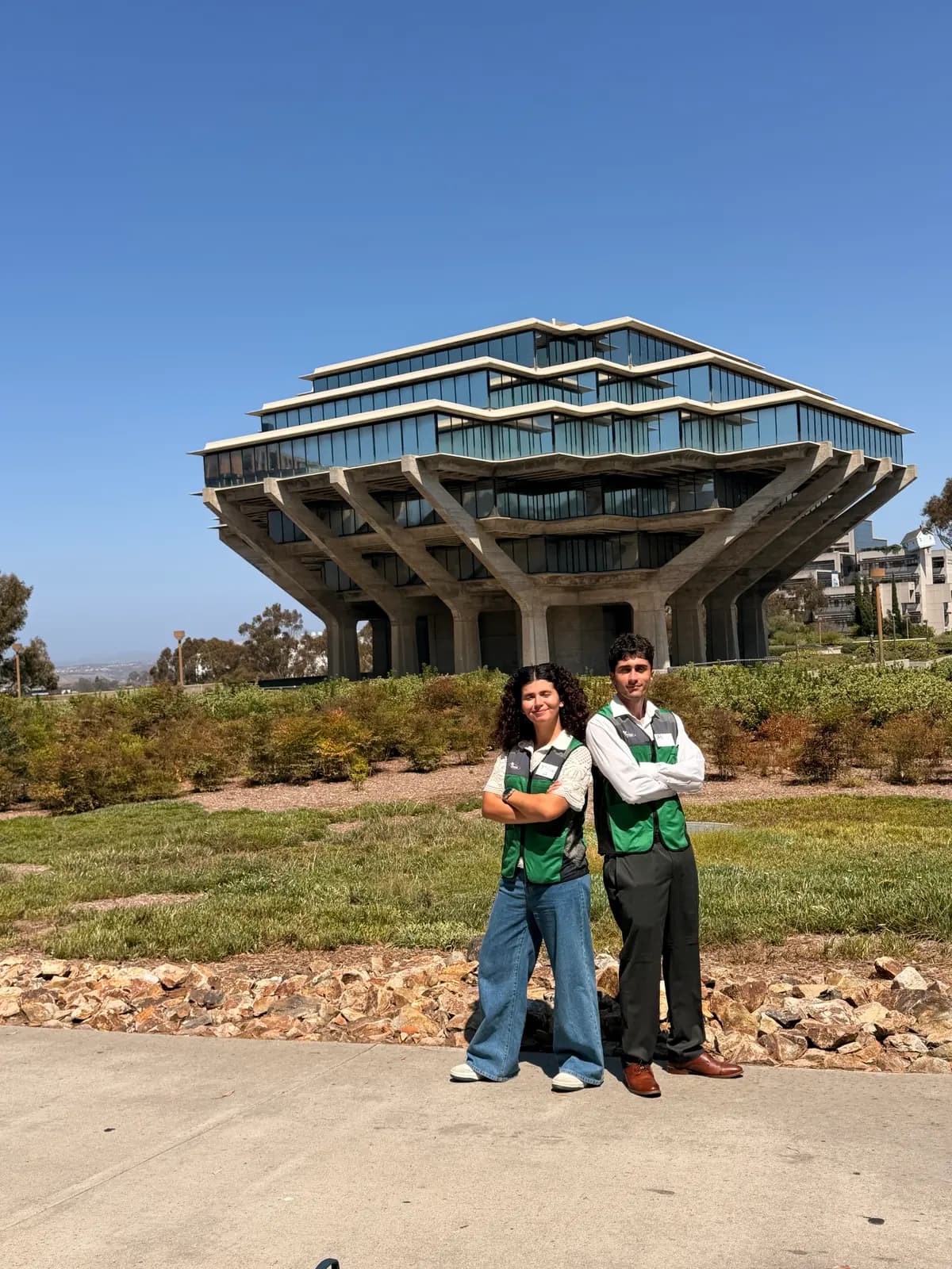 Two people in green ReDorm vests standing with arms crossed in front of the Geisel Library at UC San Diego