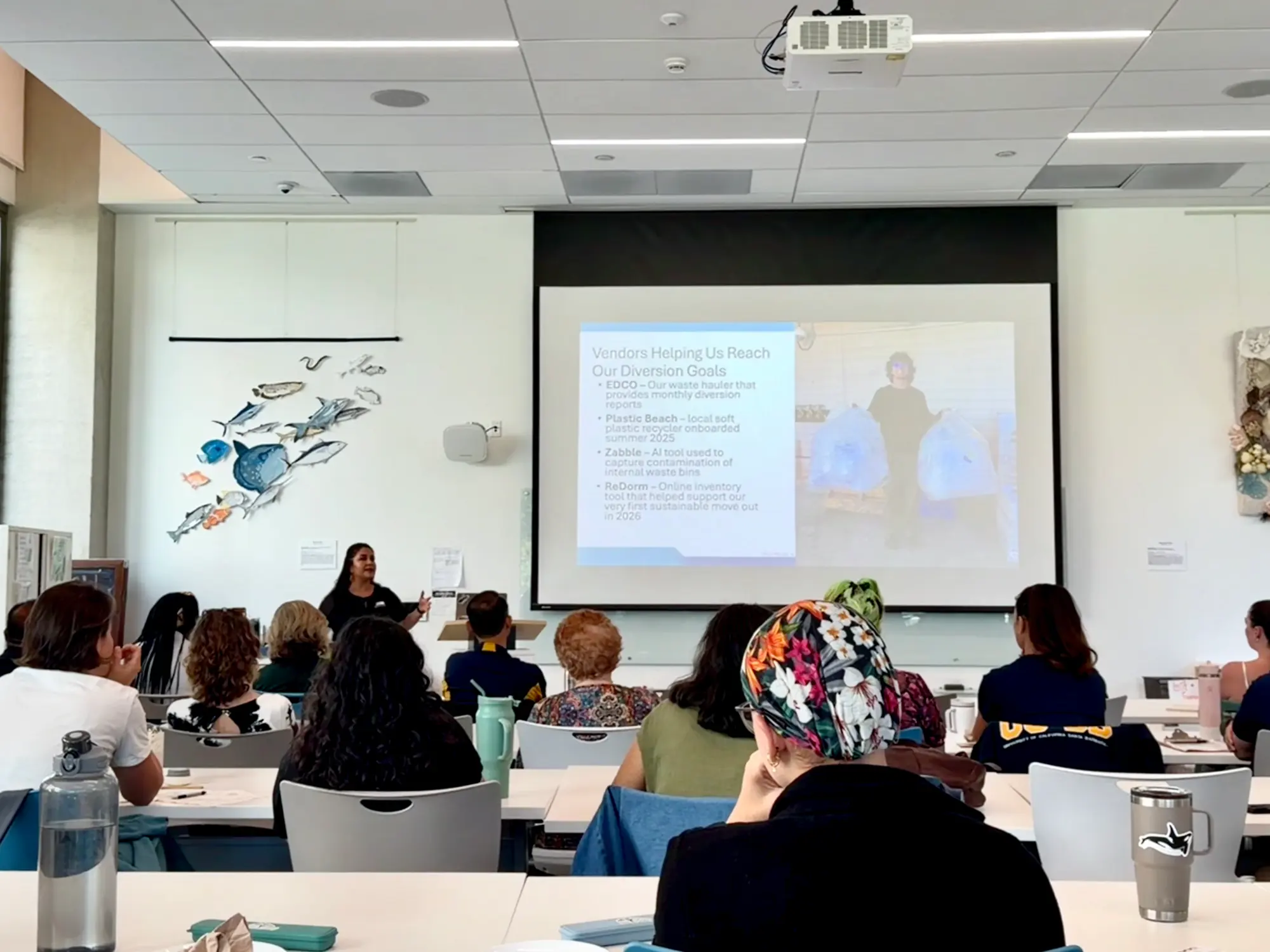 A presenter stands beside a projected slide titled Vendors Helping Us Reach Our Diversion Goals in front of a seated audience in a campus classroom